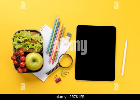 Tablet with blank screen, school supplies, stationery, and lunch box on yellow background. Education and nutrition. Flat lay with blank space for your Stock Photo