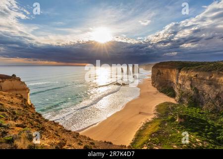 Australia, Victoria, View of sun shining through storm clouds over sandy beach in Port Campbell National Park with Twelve Apostles in background Stock Photo