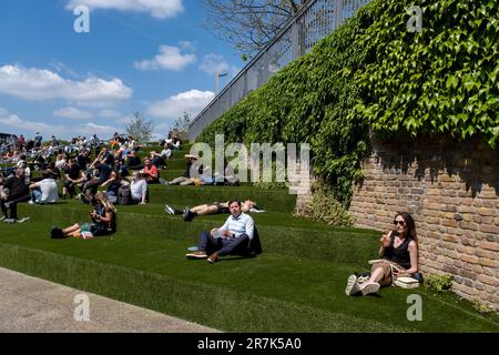 People relax in the Spring sunshine on the steps covered in fake grass leading up to Granary Square in Kings Cross on 24th May 2023 in London, United Kingdom. Kings Cross Central is a multi-billion pound mixed-use development on a site owned and controlled by the Kings Cross Central Limited Partnership covering approximately 67 acres of former railway land. King’s Cross is one of the largest and transformative redevelopments seen in London in recent times, changing what was an industrial site into a rejuvenated and vibrant area with squares, parks, homes, shops, offices, galleries, bars, resta Stock Photo