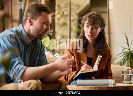 Young psychologist talking to patient and making notes in notepad during their meeting in office Stock Photo