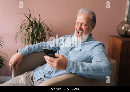 Medium shot of laughing senior aged male holding smartphone using mobile online app, cheerful looking to screen sitting on armchair. Stock Photo