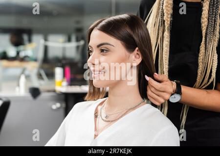 hairdresser and happy female client in beauty salon, beauty worker with braids standing near tattooed woman, discussing hair treatment, hair extension Stock Photo