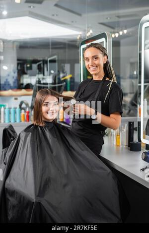 salon services, hairdresser with round brush styling hair of female customer, happy brunette woman with short hair, beauty salon, hair volume, looking Stock Photo