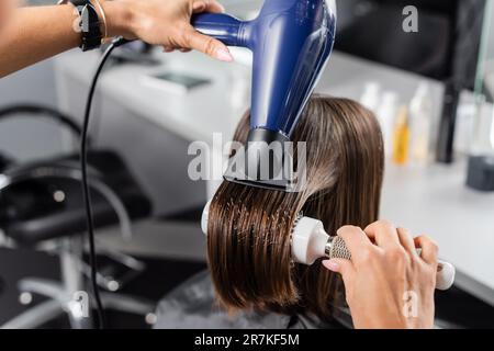 salon blow dry, professional hairdresser with round brush and hair dryer styling hair of female customer, brunette woman with short hair, beauty salon Stock Photo