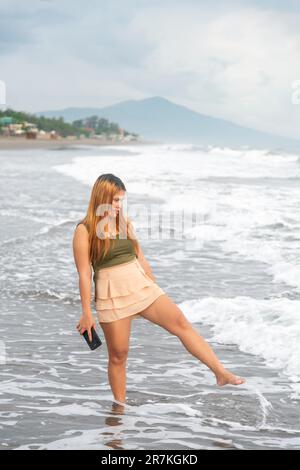 Holding her phone for selfies,on a beach near Manila at dusk,aimlessly strolling the warm shoreline waters,along smooth sands,having fun,dancing aroun Stock Photo