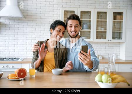 Cheerful loving couple of different nationalities, talking via video conference using smart phone, during a morning breakfast, looking into the camera of the mobile phone, smiling happily Stock Photo