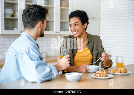 Happy young family couple, caucasian man and african american woman, having breakfast at cozy home kitchen with healthy porridge, happy to spend time together at weekend morning, communicating, smile Stock Photo
