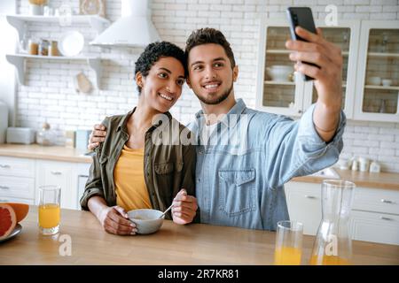 Happy beautiful couple in love, of different nationalities, bloggers, using smartphone during a morning breakfast, taking a selfie photo, looking into the camera of the mobile phone, smiling happily Stock Photo