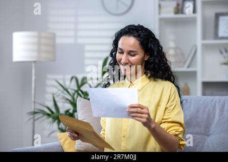 Joyful and happy woman at home received mail envelope with notification, hispanic woman reading good news on paper and happy sitting on sofa in living room . Stock Photo