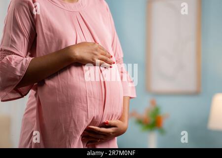 Close up shot of Pregnant woman standing by touching or feeling tummy at home - concept of expecting baby, motherhood and Emotional connection. Stock Photo
