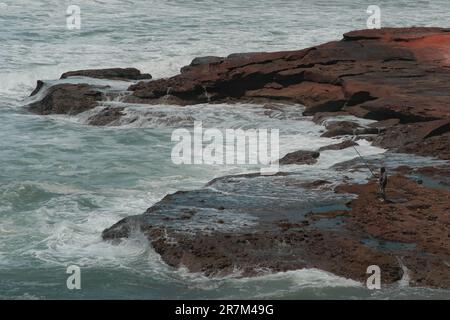 Man holds fishing rod to catch fish amid wild waves in Legzira Beach. Person stands on sandstone rock formation. Rugged landscape in Tiznit Province. Stock Photo