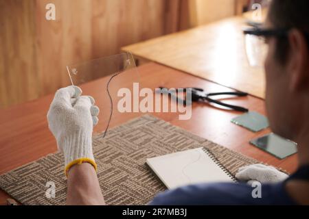 Rear view of craftsman in gloves holding piece of glass sitting at his workplace, he creating artwork from glass Stock Photo