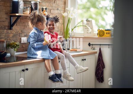 Two adorable little girls sitting on kitchen counter top, enjoying a snack together, eating a watermelon and a muffin. Home, lifestyle, family concept Stock Photo