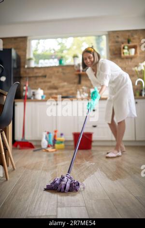 Joyful housewife cleaning the kitchen, wet mopping the floor, with chemical bottles in the background. Hygiene, home, lifestyle concept. Stock Photo