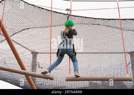 Teenage teen girl in climbing harness equipment, green sports safety helmet. Rope amusement park. Fastening attaching carabiner to safety rope. Hangin Stock Photo