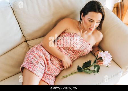 Beautiful stylish woman lying on on white leather couch, wearing trendy pink tweed suit, holding tender peony flower Stock Photo