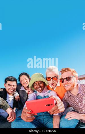 Vertical portrait of group of multiracial happy friends having fun using a cellphone app to browse on internet. Young excited people smiling and taking a selfie photo together with a smar tphone. High quality photo Stock Photo