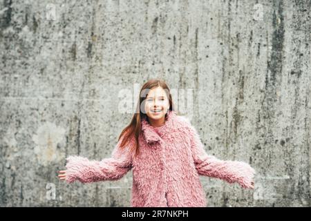 Outdoor portrait of excited little girl wearing faux fur pink coat, posing against grey city wall, arms wide open Stock Photo