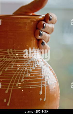 painted with white enamel earthenware jug in female hands. ceramic dishes on a blurred background close up. vertical. Stock Photo