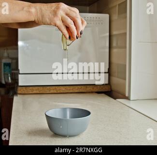 female hands breaking a chicken egg, pouring out a bowl against the background of a kitchen interior. baking, cooking. Stock Photo