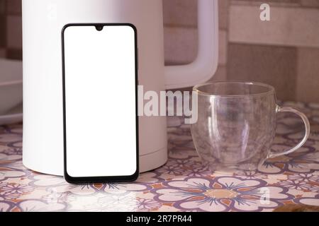 a phone with a blank white screen stands near a white teapot and a cup on the kitchen table Stock Photo