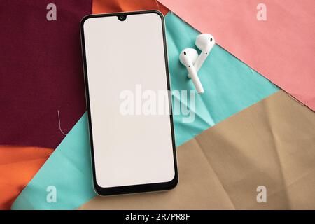 a phone with a white screen and wireless headphones lies on lies on multi-colored fabrics close-up Stock Photo