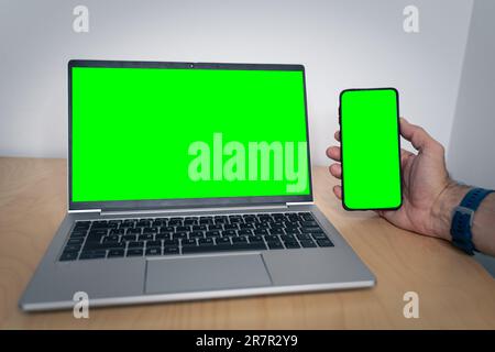 Mockup image of a man holding a mobile phone with green screen and a laptop with blank green screen on table Stock Photo