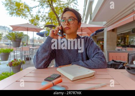 A young woman in her twenties is sitting at a cafe table with a cell phone in her hand, seemingly engaged in a phone conversation Stock Photo