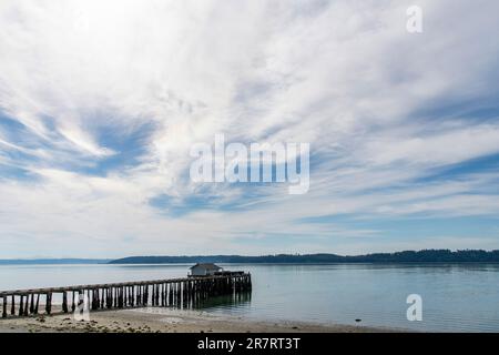 Panoramic view over the water of Penn Cove near Coupeville, WA, USA with a wooden fishing dock and building extended in the water against a blue sky w Stock Photo