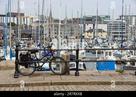 A single black bicycle rests againsts the railings on the cobbled West Pier on Plymouth’s Barbican. In soft focus beyond the inner basin with yachts a Stock Photo