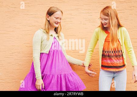 Outdoor portrait of two funny preteen girls, wearing colorful clothes, posing next to orange background Stock Photo