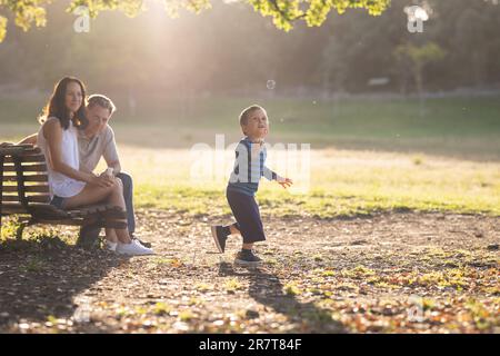 Cute white family in the park - a little boy chasing soap bubbles and his parents watching him. Mid shot Stock Photo