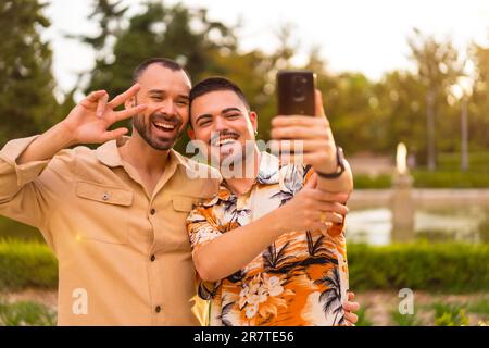 Homosexual boyfriends making a video call laughing at sunset in a park in the city. Diversity and lgbt concept Stock Photo