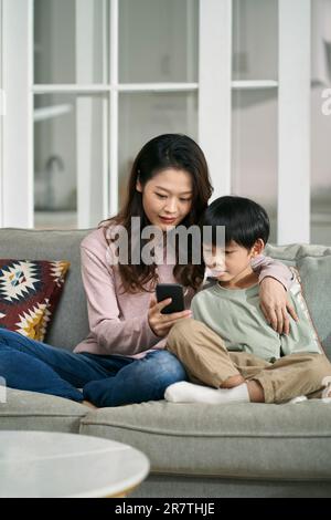 happy asian mother and son sitting on family couch looking at cellphone photos together Stock Photo