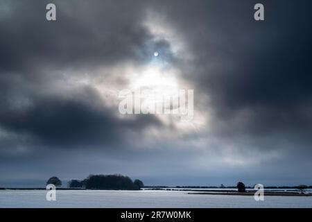 Sunlight through dark cloudy winter sky and woodland landscape in wintertime in Swinbrook in The Cotswolds, Oxfordshire Stock Photo