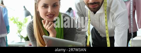 Man and woman designers are discussing new collection of clothes on digital tablet in workshop. Workers of online fashion store in modern warehouse. Stock Photo