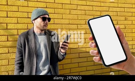 Mobile phone with blank screen in hand, urban stylish man standing next to brick wall in city and using his smart phone. Stock Photo