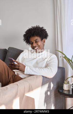 Happy gen z African American teen sitting on couch using mobile phone. Stock Photo
