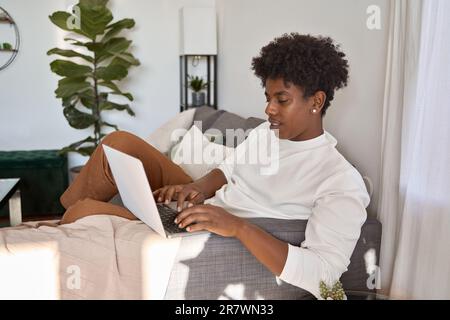 African American guy student sitting on couch using laptop computer at home. Stock Photo