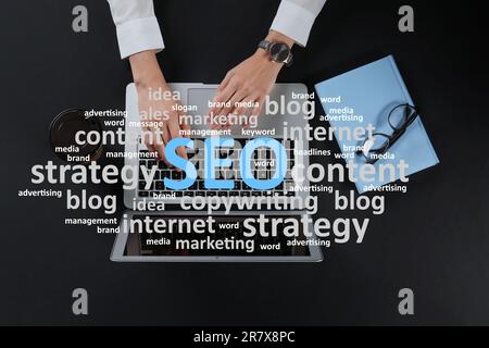 Search engine optimization (SEO). Cloud of words above copywriter with laptop at black table, top view Stock Photo