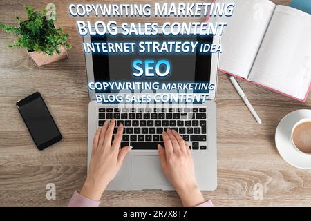 Search engine optimization (SEO). Copywriter working on laptop at wooden table, top view. Many words coming out of computer Stock Photo