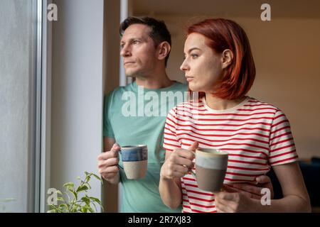 Upset girl and man hold cup of coffee, look out window, feeling sad and anxious. Family problems Stock Photo