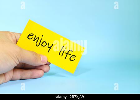 Enjoy life reminder concept. Hand holding a bright yellow paper message note. Stock Photo