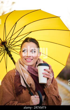 Caucasian young woman with coffee and yellow umbrella smiling in autumn. Stock Photo