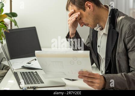 Frustrated overworked caucasian businessman, entrepreneur, product manager, programmer, sits at a desk in the office, massaging the bridge of his nose Stock Photo