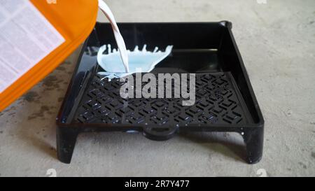 A man pours paint into a drawing tray. Close-up of white paint being poured into a black paint roller tray. White paint is poured into a black tray. Stock Photo