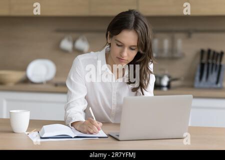 Young woman writing notes, sitting at kitchen table with laptop Stock Photo