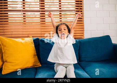 Happy cute girl with hands up sitting on comfortable sofa Stock Photo