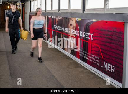 Young couple walking past an advertising hoarding for London North Eastern Railway (LNER) for smarter travel and book direct Stock Photo