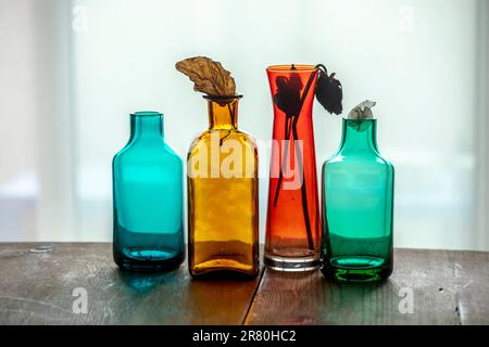 Still life of colored glass bottles and vases with dried flowers on a wooden table in front of a window with white curtains and soft light Stock Photo